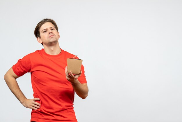 Front view of ambitious young guy in red blouse holding small box and posing for camera on white background