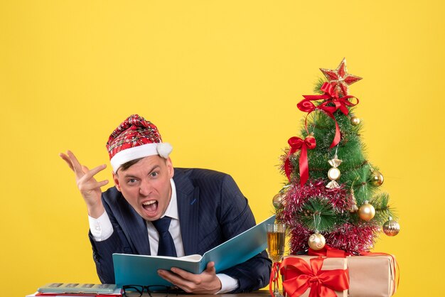 Front view of angry business man with santa hat sitting at the table near xmas tree and presents on yellow wall