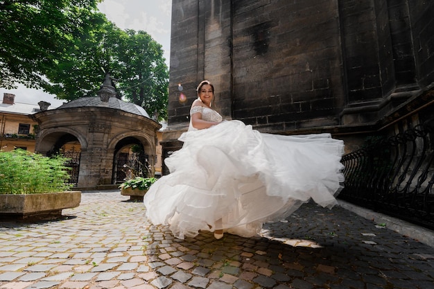 Free photo front view of attractive bride in luxurious dress waving her skirt smiling and looking at camera while walking near ancient buildings during sunny wedding day xa