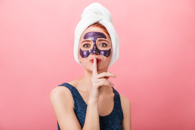 Front view of attractive woman showing secret sign while doing spa treatment. Studio shot of pretty girl with face mask isolated on pink background.