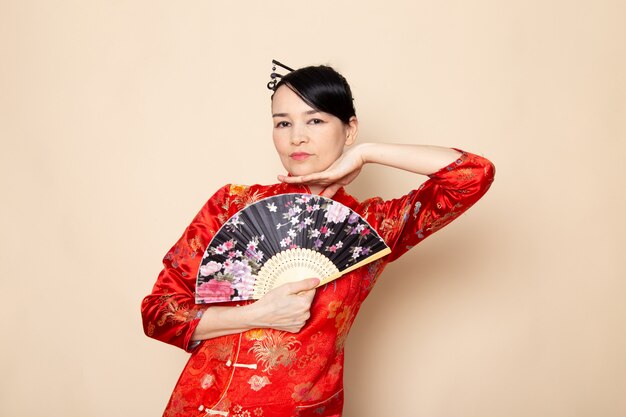 A front view beautiful japanese geisha in traditional red japanese dress with hair sticks posing holding folding fan elegant on the cream background ceremony japan