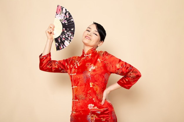 Free Photo a front view beautiful japanese geisha in traditional red japanese dress with hair sticks posing holding folding fan smiling on the cream background ceremony japan