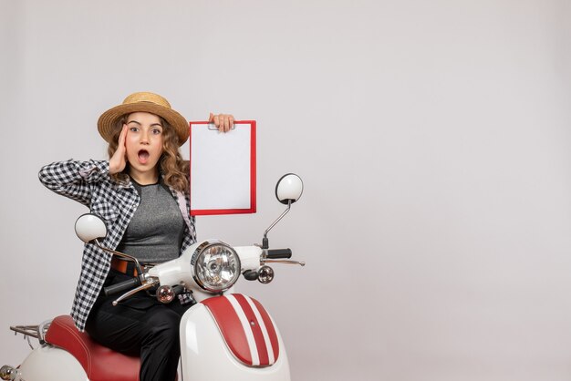 Front view of bewildered young girl on moped holding up clipboard on grey wall