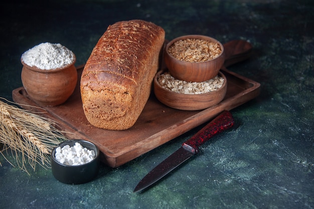 Front view of black bread slices flour in a bowl on wooden board and knive spikes raw oatmeal wheat on mixed colors distressed background