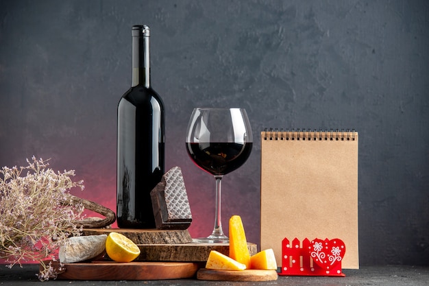 Front view black wine bottle wine in glass cheese cut lemon a piece of dark chocolate on wooden boards dried flower branch notepad on red table