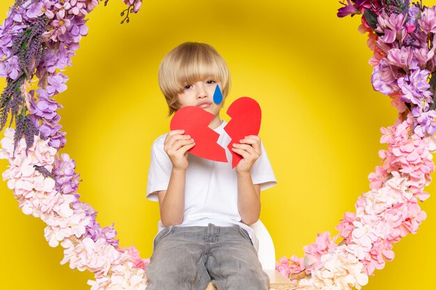 Free photo a front view blonde haired boy in white t-shirt with heart shape sitting on the flower made stand on the yellow floor