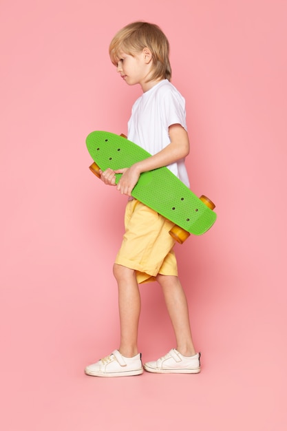 A front view blonde haired boy in white t-shirt and yellow jeans holding green skateboard on the pink space