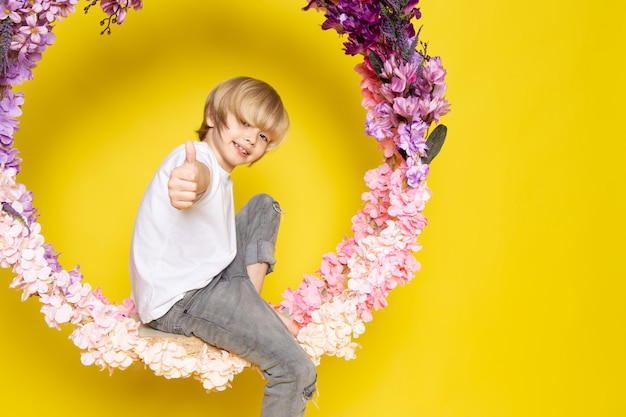 A front view blonde smiling child in white t-shirt sitting on the flower made stand on the yellow desk