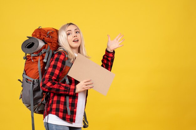 Front view blonde traveler woman with her backpack holding cardboard waving hand standing on yellow wall