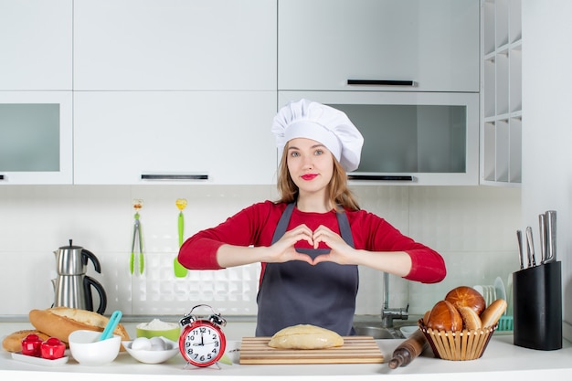 Front view blonde woman in cook hat and apron making heart sign in the kitchen