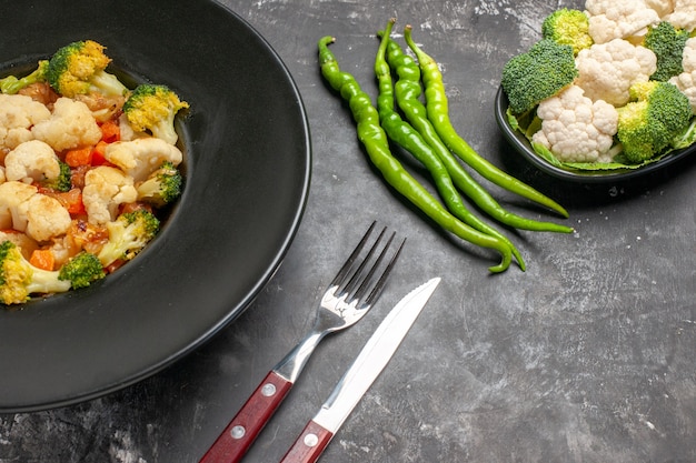 Front view broccoli and cauliflower salad on black oval plate fork and knife green hot peppers on dark background