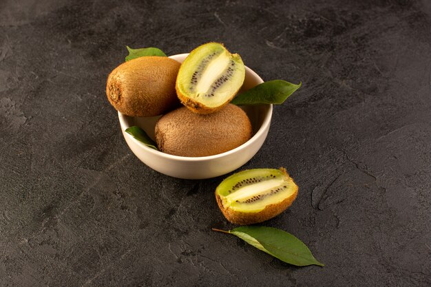 A front view brown kiwis fresh ripe isolated juicy mellow whole and sliced fruits along with green leaves inside white plate on the dark background exotic fruit fresh