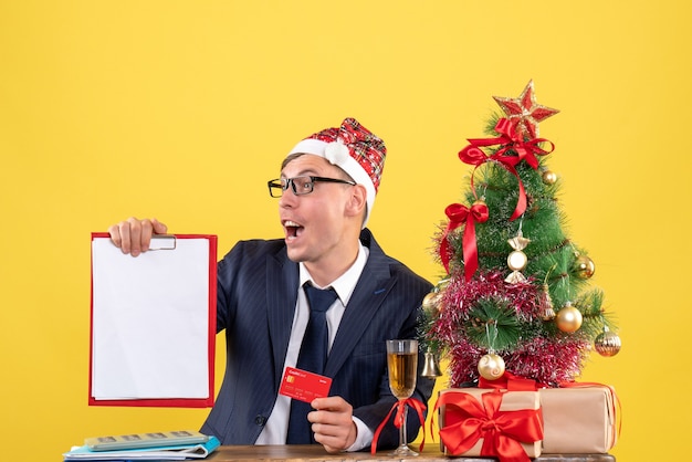 Front view of business man holding clipboard and card sitting at the table near xmas tree and presents on yellow