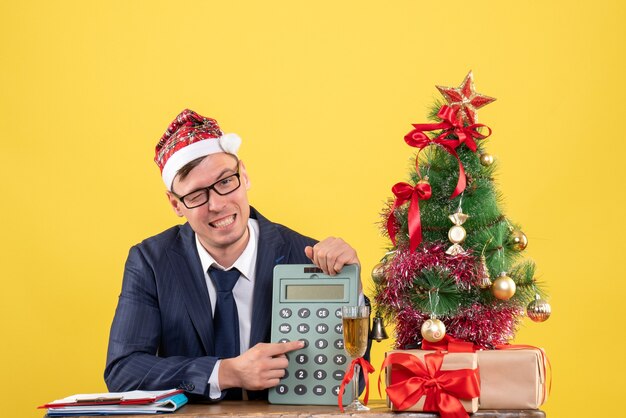 Front view of business man showing calculator sitting at the table near xmas tree and presents on yellow