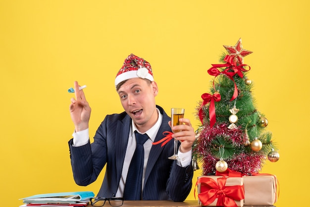 Front view of business man toasting holding noisemaker sitting at the table near xmas tree and presents on yellow