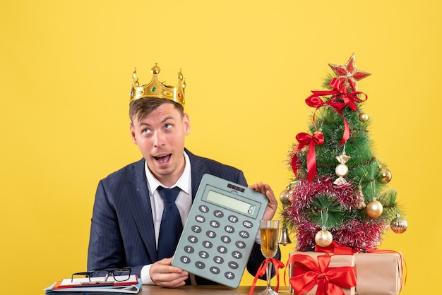 Free photo front view of business man with crown holding calculator sitting at the table near xmas tree and presents on yellow