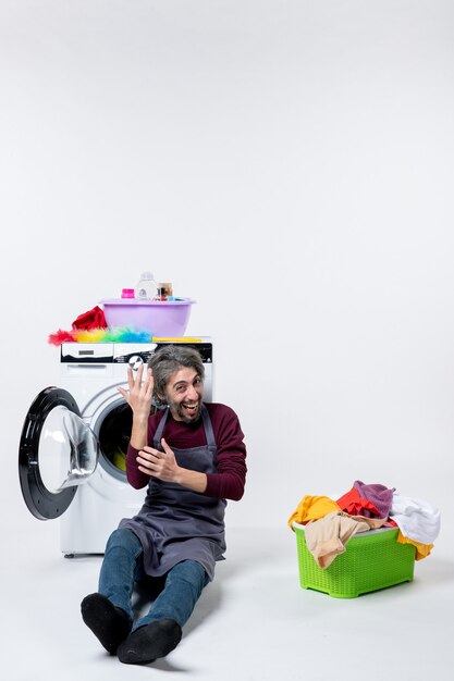 Front view cheery young housekeeper sitting in front of washer laundry basket on white background