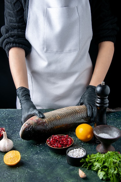 Free photo front view chef in apron putting raw fish on chopping board pepper grinder pomegranate seeds in bowl on table