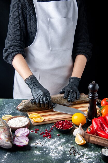 Front view chef with black gloves chopping raw fish on wood board pepper grinder flour bowl pomegranate seeds in bowl on kitchen table