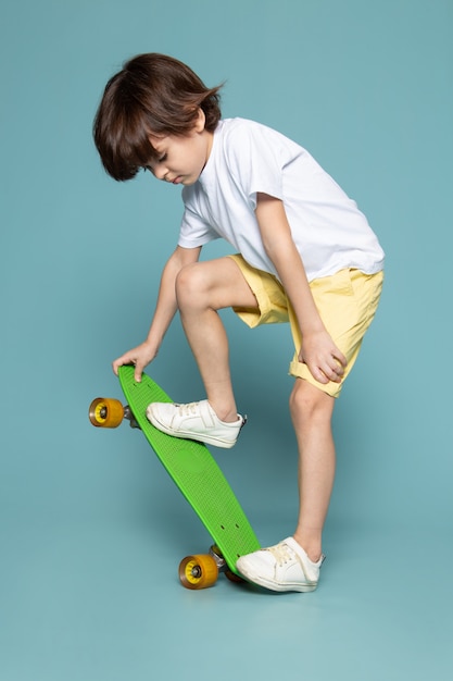 A front view child boy in white t-shirt and yellow jeans riding green skateboard on the blue space