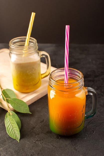 A front view cold cocktails colored inside glass cans with colorful straws green leaves on the wooden cream desk and dark