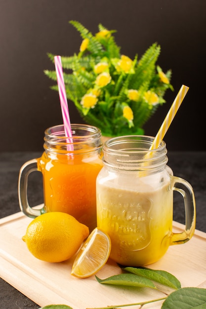 A front view cold cocktails colored inside glass cans with colorful straws lemons green leaves flowers on the wooden cream desk and dark