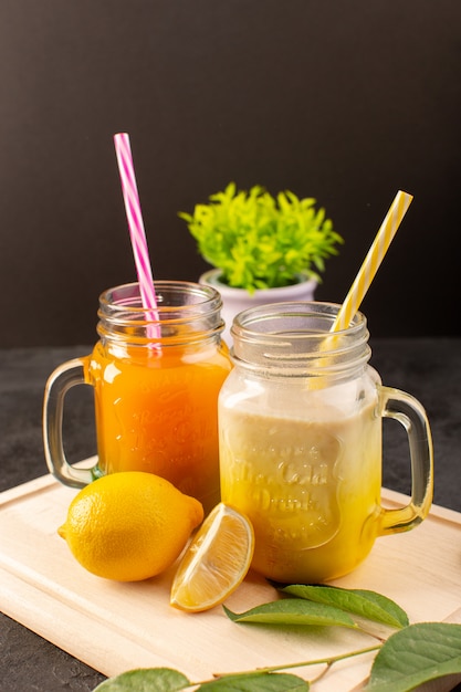 A front view cold cocktails colored inside glass cans with colorful straws lemons green leaves on the wooden cream desk and dark