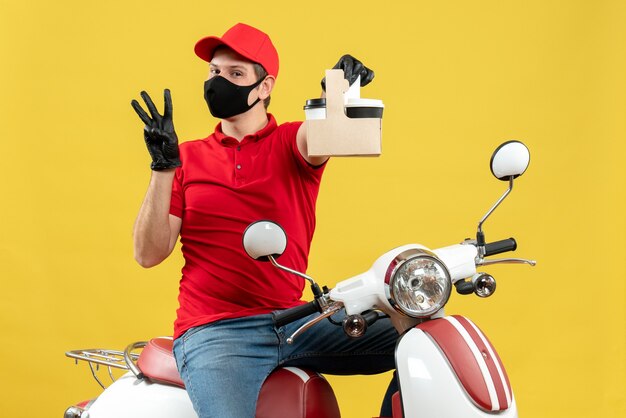 Front view of confident courier man wearing red blouse and hat gloves in medical mask sitting on scooter holding orders showing three