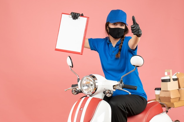 Free photo front view of confident courier woman wearing medical mask and gloves sitting on scooter holding empty paper sheets delivering orders making ok gesture on pastel peach background