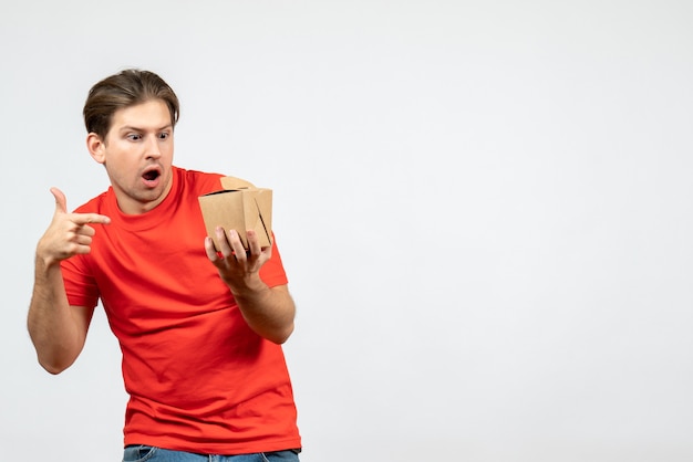 Front view of confused emotional young guy in red blouse pointing small box on white background