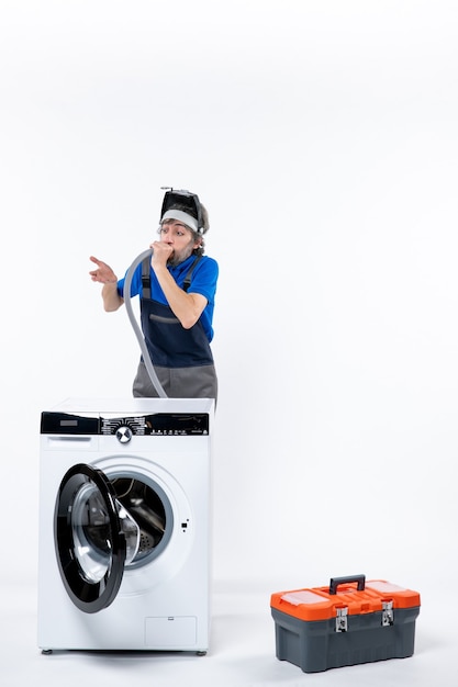 Front view of confused repairman in uniform standing behind washing machine blowing out pipe on white wall