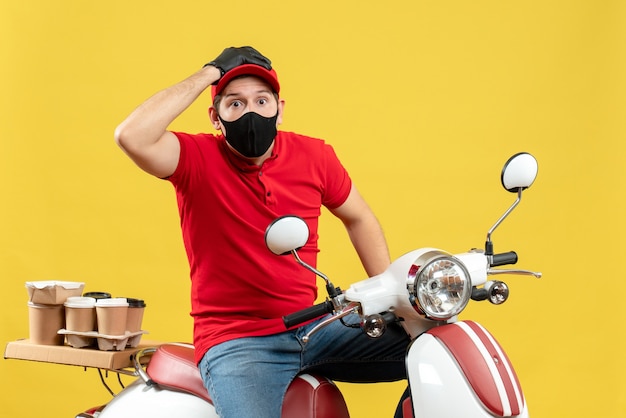 Front view of confused young adult wearing red blouse and hat gloves in medical mask delivering order sitting on scooter on yellow background