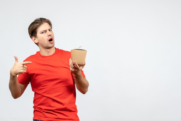 Front view of confused young guy in red blouse pointing small box on white background