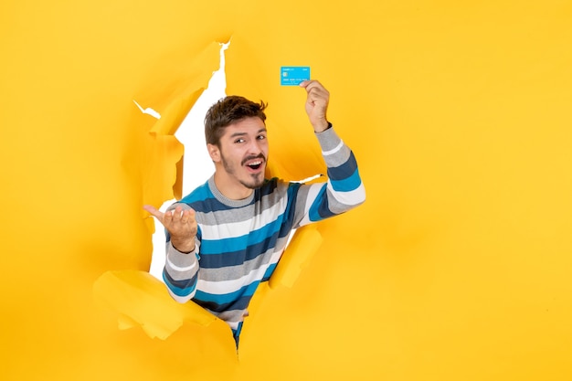 Free Photo front view confused young man holding up card looking through torn paper yellow wall