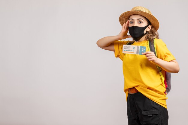 Front view of confused young woman with black mask holding travel ticket on white wall