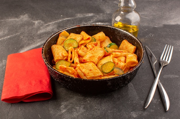 A front view cooked italian pasta with tomato sauce and cucumber inside pan on the dark surface