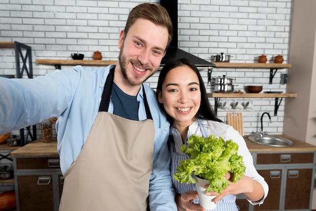 Front view of couple capturing selfie with woman holding fresh green lettuce in kitchen