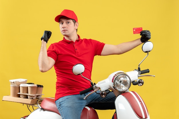 Front view of courier man wearing red blouse and hat gloves in medical mask delivering order sitting on scooter pointing bank card showing back