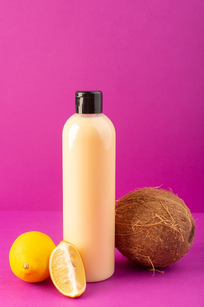 A front view cream colored bottle plastic shampoo can with black cap along with lemons and coconut isolated on the purple background cosmetics beauty hair