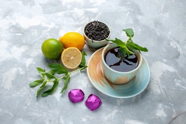 Front view cup of tea with fresh lemons candies and dried tea on light table, tea fruit citrus color