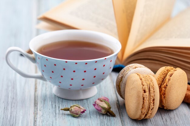 Front view of cup of tea with macarons and an open book on a gray surface