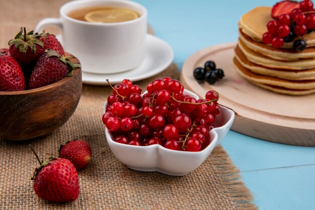 Front view of cup of tea with a slice of lemon and pancakes with red and black currants and strawberries on a blue surface