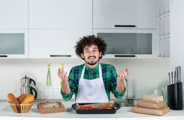 Front view of curious man standing behind table with freshly-baked pastry on it in the white kitchen