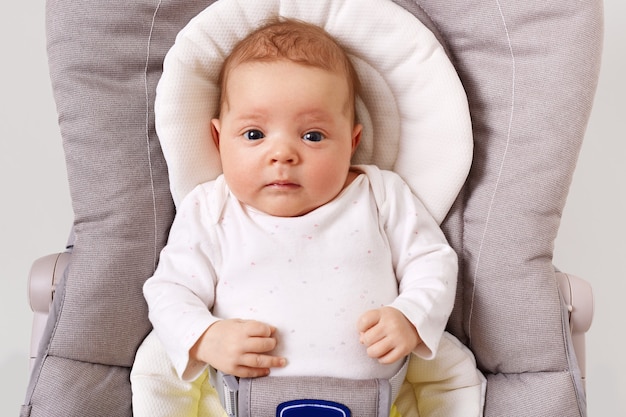 Front view of curious new born baby wearing white podysuit lying in child bouncer chair