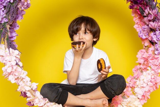 A front view cute boy eating donuts around flowers on the yellow desk