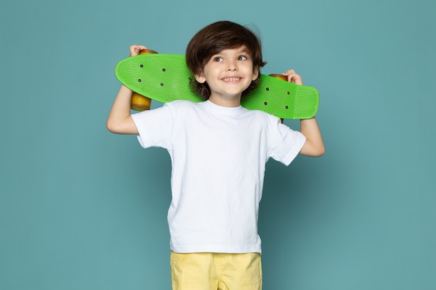 Free photo a front view cute boy in white t-shirt and yellow jeans holding green skateboard on the blue space