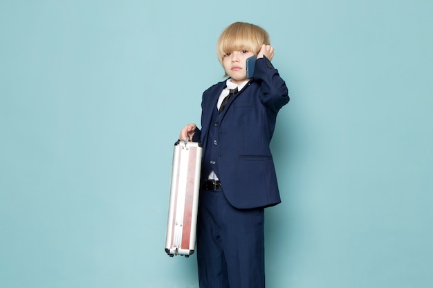 Free photo a front view cute business boy in blue classic suit posing holding brown-silver suitcase talking on the phone business work fashion