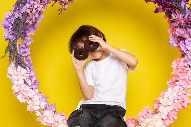 Free photo a front view cute little boy in white t-shirt holding pair of choco donuts on the yellow desk