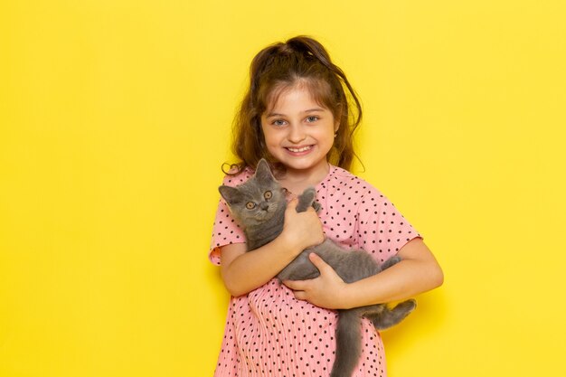 A front view cute little kid in pink dress holding grey kitten and smiling