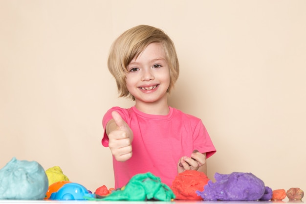 A front view cute little kid in pink t-shirt showing awesome sign playing with colorful kinetic sand